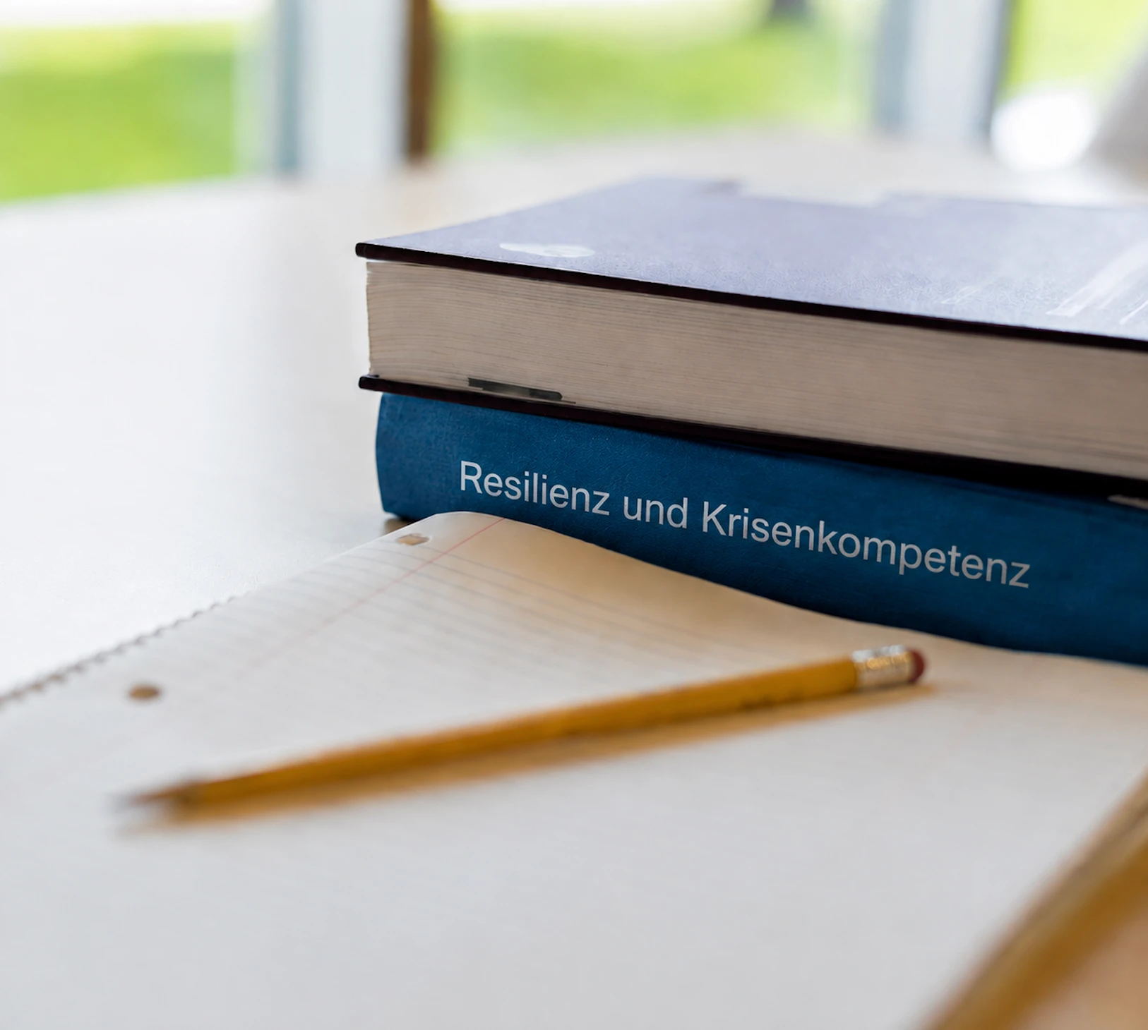 Buchrücken mit Resilienz und Krisenkompetenz auf einem Schreibtisch mit Notizbuch und Bleistift